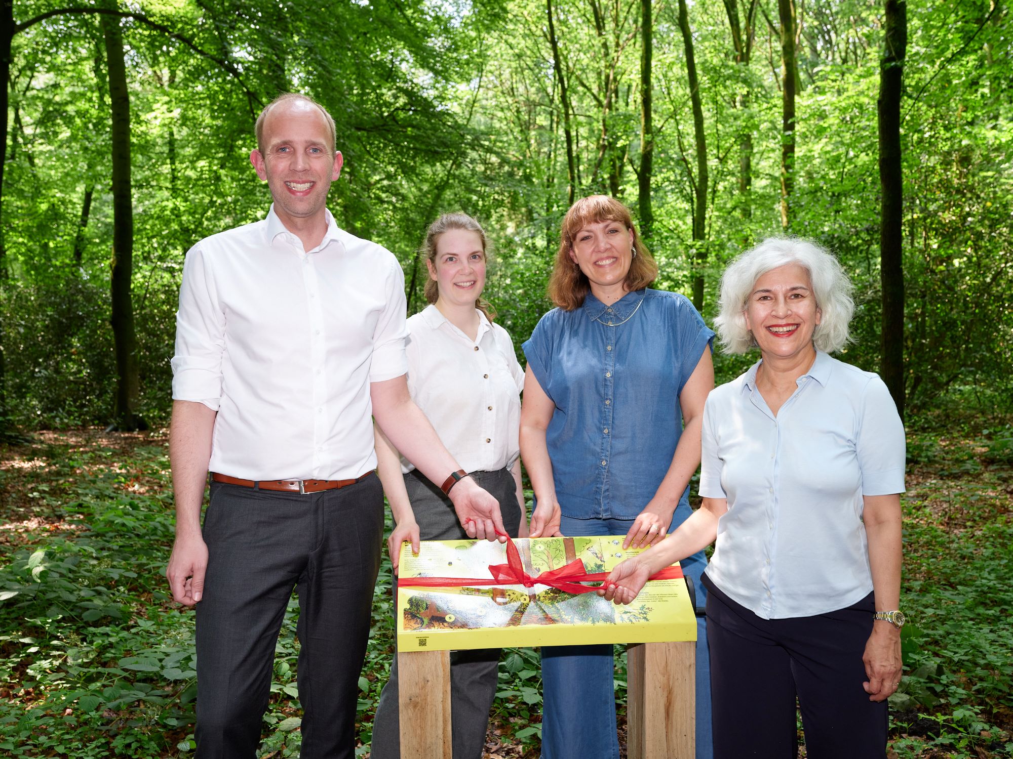 1 Mann und 3 Frauen hinter einer gelben Tafel mit Drehkurbel und roter Schleife