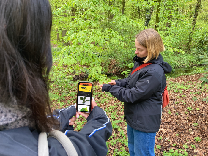 Junge Frau mit auf dem Handy geöffneter Actionbound Rallye, während eine andere junge Frau Blätter untersucht.