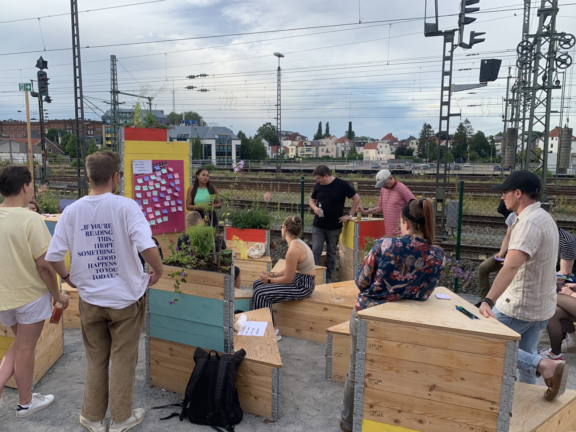 Bunte dreieckige Sitzmöbel und Hochbeete, Menschen, die Klebezettelsammelung besprechen, dahinter Gleise