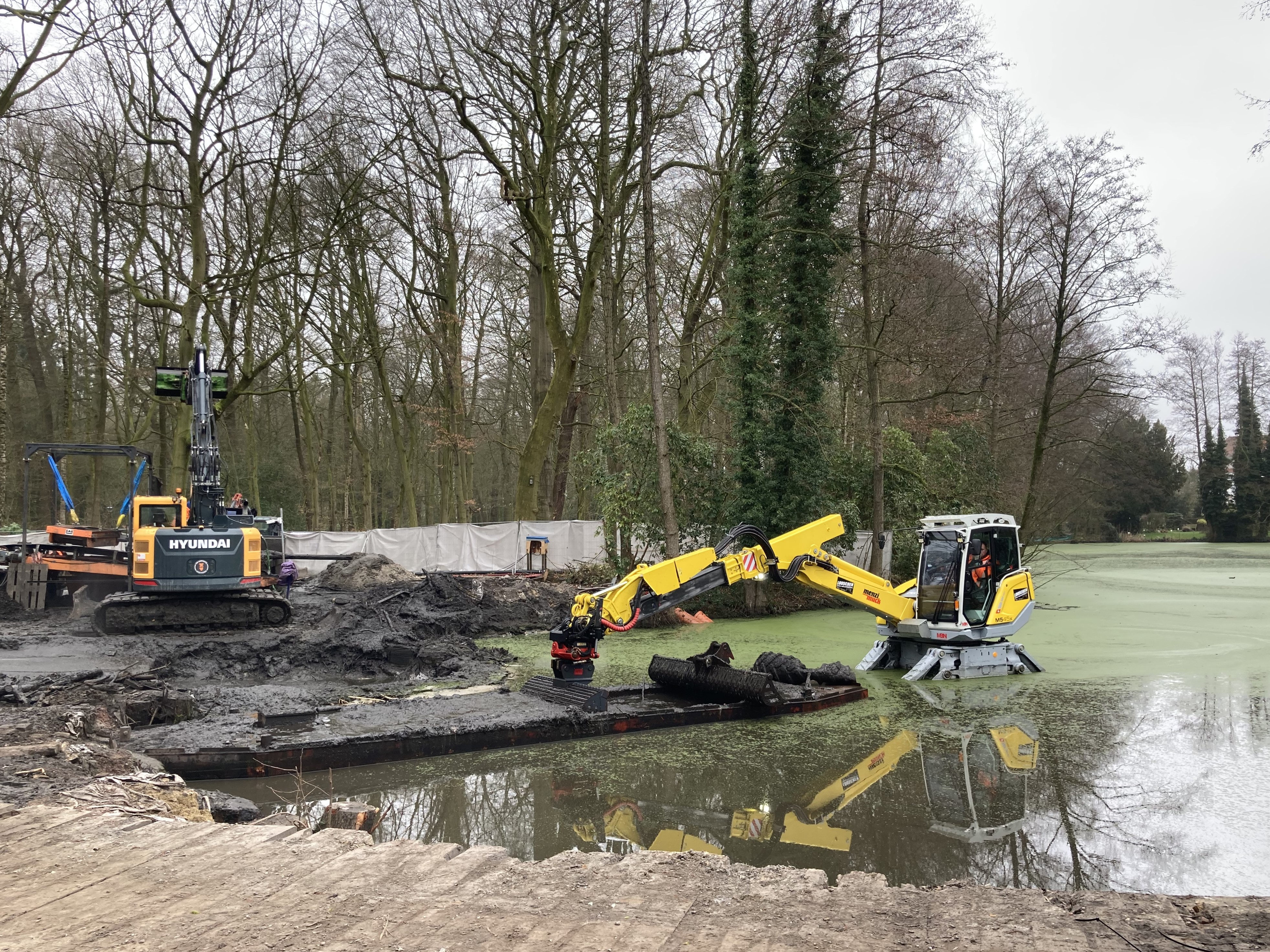 Bagger steht im Gewässer und baggert Schlamm in eine Schute