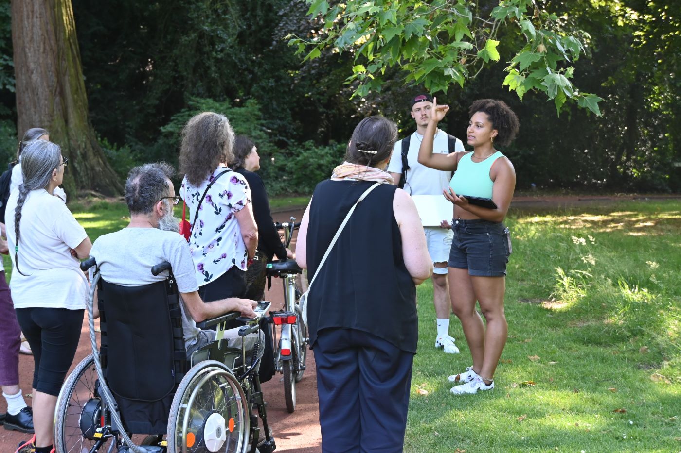 Verschiedene Menschen, einige mit Rollstuhl und Fahrrad hören Chioma Ndukwe zu, wie sie etwas über die Platane im Schlossgarten erzählt